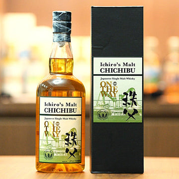 A bottle of Ichiro's Malt Chichibu Japanese Single Malt Whisky stands next to its tall black packaging box on a wooden surface, with a softly blurred bar background.